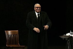 James Earl Jones standing with an overcoat on near a chair on one side and a table on the other.