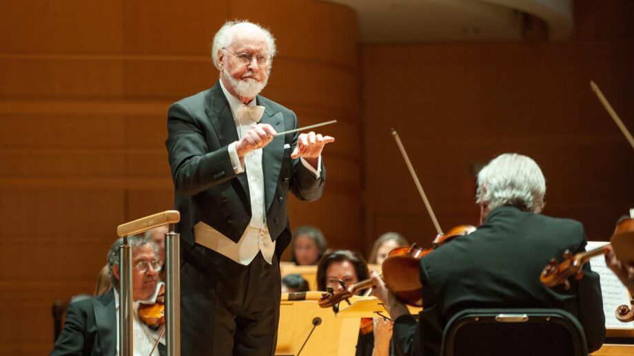 John Williams conducting, baton raised, orchestra in the background.